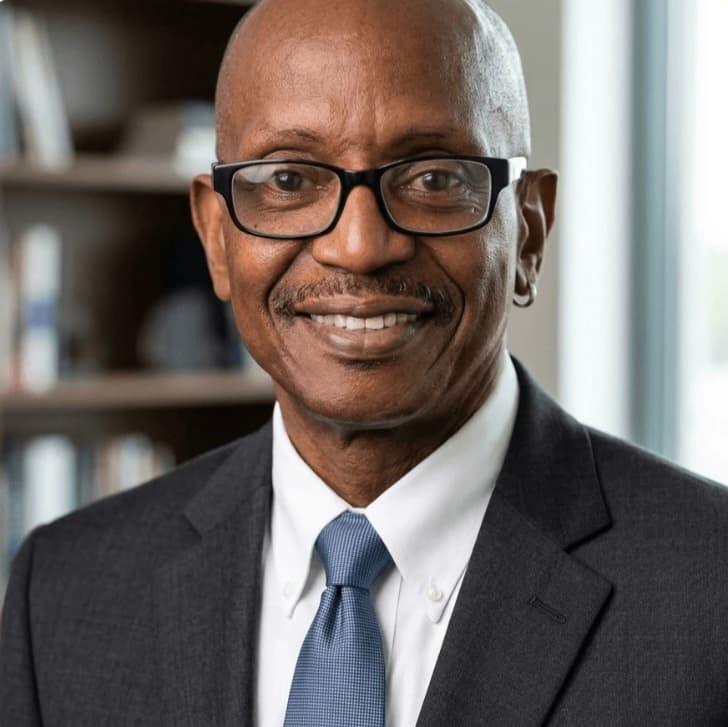 Smiling older Black man with glasses, mustache, and earring in a professional suit and tie.